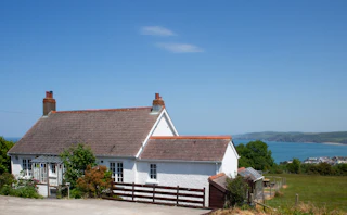 White cottage with slate roof and red chimneys beside a wooden fence, overlooking the sea